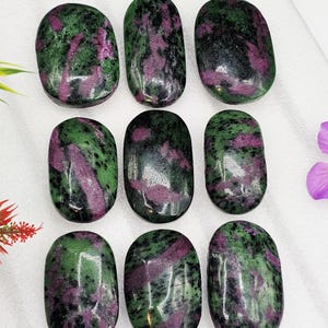 May include: Nine polished oval-shaped stones. Each stone has a mix of green and dark red colors, with a glossy finish. The stones are arranged in a grid pattern on a white surface. The overall theme is natural gemstones.