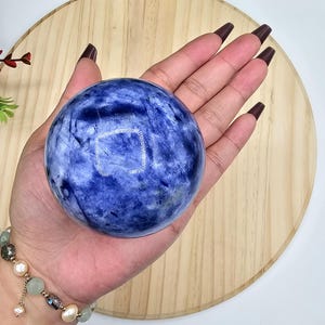 May include: A blue sodalite sphere with white veining, held in a hand. The polished crystal ball is a deep blue color with lighter blue and white marbling. A bracelet with pearls and beads is visible on the wrist.