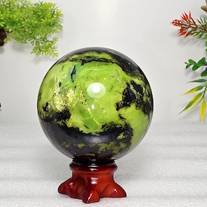May include: A polished spherical stone featuring a vibrant lime green and deep black marbled pattern. The sphere is displayed on a small, dark reddish-brown wooden stand with a tripod base, showcasing a smooth, reflective surface with natural inclusions.