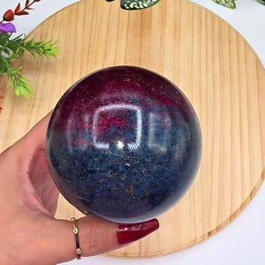 May include: A polished spherical stone featuring a distinct color split with a deep magenta-red upper section and a dark navy-blue lower section. The surface of the sphere is smooth and reflective, showing natural mineral textures and variations.
