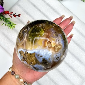 May include: A polished gemstone sphere with a mix of colors including brown, yellow, green, and white. The sphere has a natural, organic appearance with visible patterns and textures. The gemstone is held in a hand, showcasing its size and detail.