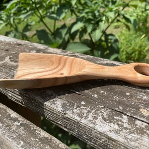 May include: A wooden scoop with a handle that has a ring at the end. The scoop is made of light brown wood with a natural finish.