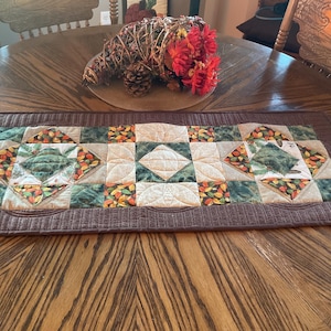 May include: A brown quilted table runner with a patchwork design featuring green, brown, and cream squares with a fall leaf pattern. The runner is on a wooden table.