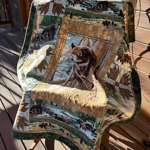 May include: A quilted blanket featuring a bear theme. The blanket has a central image of a bear in a forest setting, surrounded by panels with bear and forest imagery. The border is green, and the overall color palette includes browns, greens, and tans.