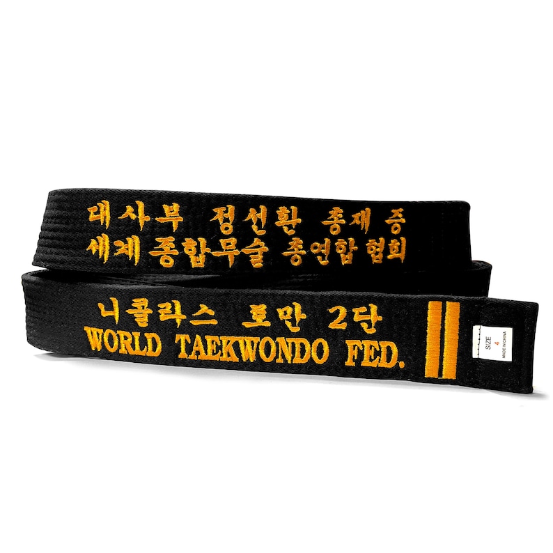 May include: Black Taekwondo belt with gold embroidered text. The belt displays Korean characters and the words "WORLD TAEKWONDO FED." and "NICHOLAS TOMAN 2 DAN". The belt has two vertical gold stripes and a size 4 label.