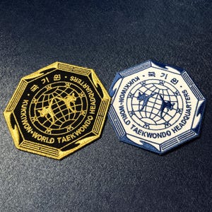 Puede incluir: Dos parches bordados con formas octogonales. Uno es negro y dorado, el otro blanco y azul. Ambos presentan un diseño de globo terráqueo con figuras de Taekwondo y el texto "Kukkiwon World Taekwondo Headquarters".