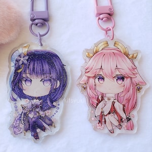 Puede incluir: Dos llaveros de personajes de estilo anime con detalles de purpurina. El llavero de la izquierda presenta un personaje de cabello morado con una flor blanca en el cabello. El llavero de la derecha presenta un personaje de cabello rosa con una corona dorada. Ambos llaveros tienen un fondo transparente con flores de cerezo rosas.