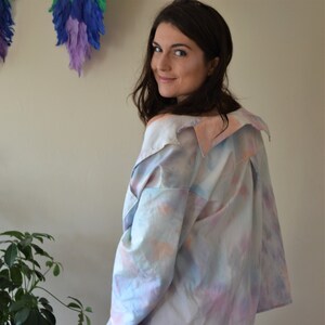 Può includere: Una donna indossa una camicia tie-dye blu chiaro, rosa e bianca con una vestibilità ampia e maniche lunghe. La camicia ha un colletto che è ripiegato verso il basso. La donna indossa anche jeans patchwork blu.