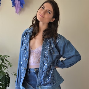 Peut inclure: Une veste en jean bleu avec des embellissements en sequins sur le devant. La veste est déboutonnée et portée sur un caraco en dentelle de couleur lilas clair. La personne portant la veste porte également un pantalon en jean bleu.