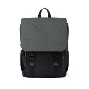 May include: A black backpack featuring a dark grey top flap decorated with a repeating black line art pattern of various shark teeth. The bag includes a top handle, two vertical straps with black plastic buckles, and side pockets for storage.