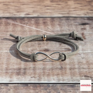 May include: A grey suede bracelet with a silver infinity symbol charm and a gold bead. The bracelet is tied with a knot at each end.