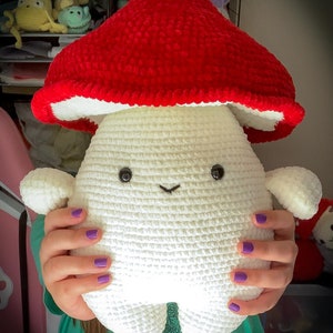 May include: A crocheted plush toy shaped like a mushroom with a white body and a red cap. The toy has a smiling face with black eyes and a small mouth.