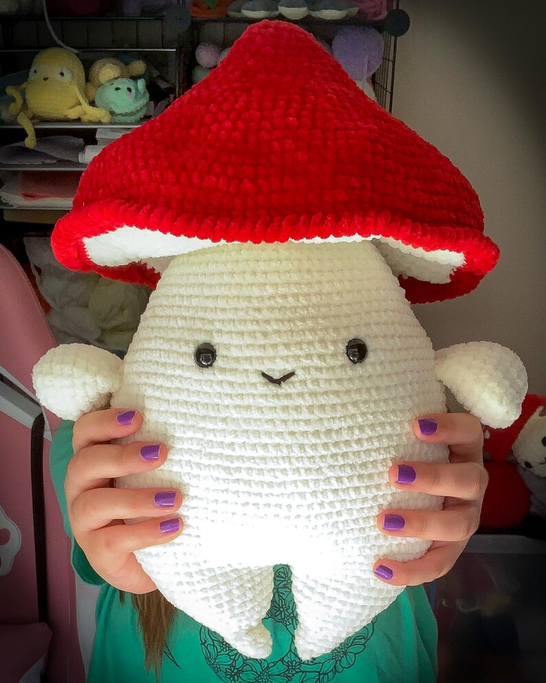 May include: A crocheted plush toy shaped like a mushroom with a white body and a red cap. The toy has a smiling face with black eyes and a small mouth.