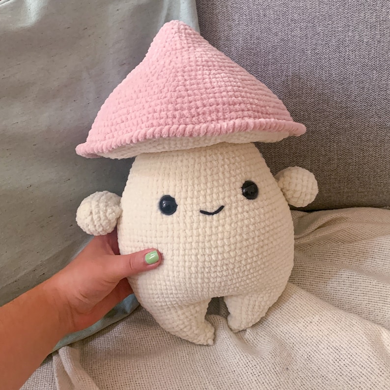 May include: A crocheted plush toy shaped like a mushroom with a pink hat and a white body. The mushroom has a smiling face with black eyes and a black mouth.