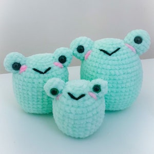 May include: Three crocheted frog plushies in different sizes, all in a light green color with black eyes and pink cheeks.