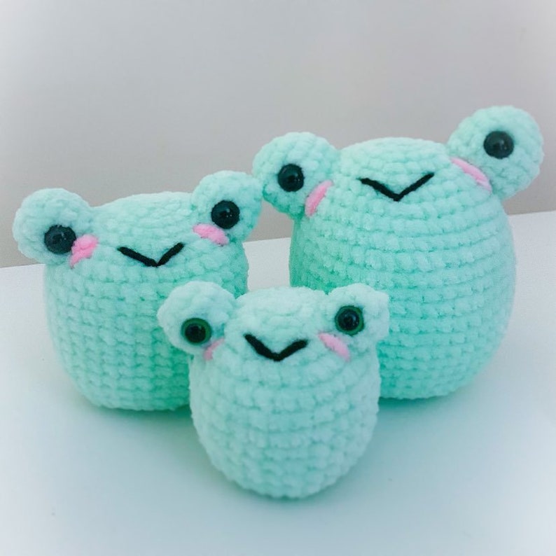 May include: Three crocheted frog plushies in different sizes, all in a light green color with black eyes and pink cheeks.