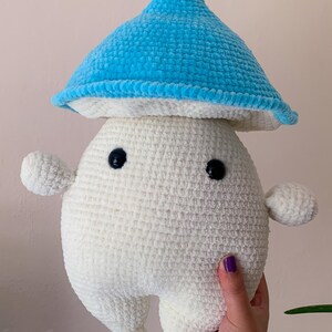 May include: A white crocheted plush toy shaped like a mushroom with a blue crocheted hat. The toy has two black eyes and a small, rounded body.