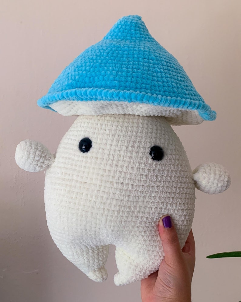 May include: A white crocheted plush toy shaped like a mushroom with a blue crocheted hat. The toy has two black eyes and a small, rounded body.