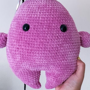 May include: A pink crocheted plush toy shaped like a blob with two black eyes. The toy has a textured surface and is made of soft yarn.