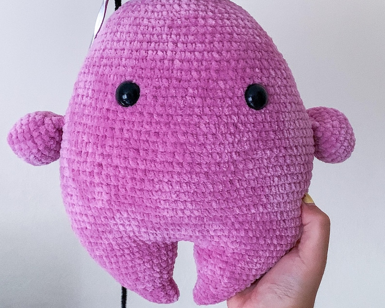 May include: A pink crocheted plush toy shaped like a blob with two black eyes. The toy has a textured surface and is made of soft yarn.