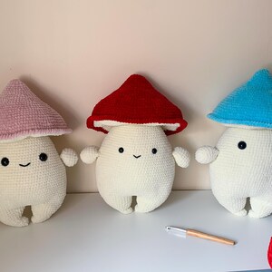 May include: Three crocheted mushroom plushies with smiling faces. The mushrooms are white with different colored tops: pink, red, and blue.
