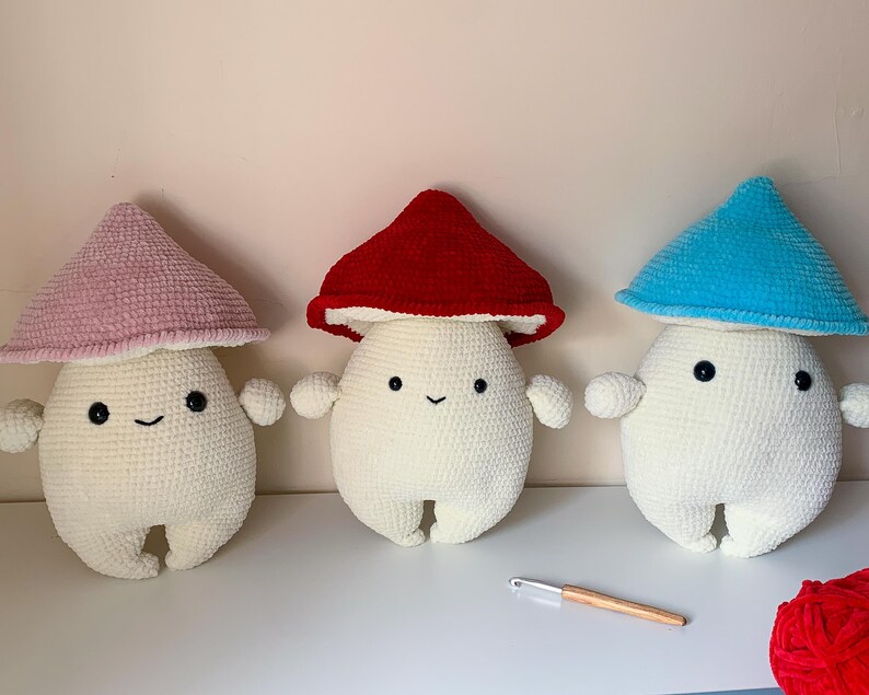May include: Three crocheted mushroom plushies with smiling faces. The mushrooms are white with different colored tops: pink, red, and blue.