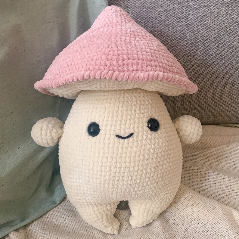 May include: A crocheted mushroom plush toy with a pink hat and a white body. The toy has a smiling face with black eyes and a small mouth.