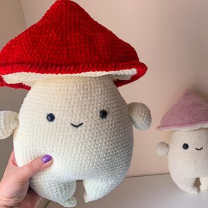 May include: Two crocheted mushroom plushies. The larger mushroom is red and white, and the smaller mushroom is pink and white. Both mushrooms have smiling faces.