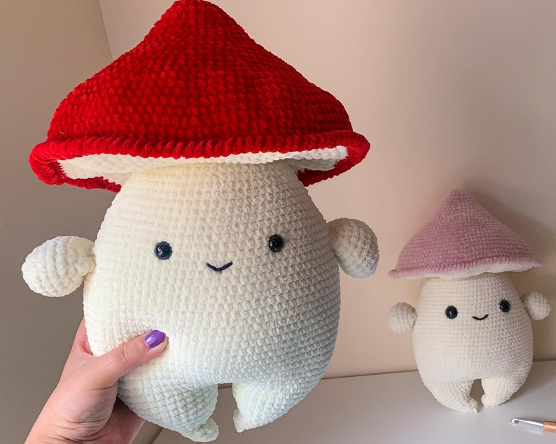 May include: Two crocheted mushroom plushies. The larger mushroom is red and white, and the smaller mushroom is pink and white. Both mushrooms have smiling faces.