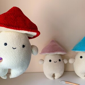 May include: Three crocheted mushroom plushies with white bodies and different colored hats. The hats are red, pink, and blue.