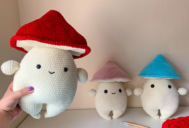 May include: Three crocheted mushroom plushies with white bodies and different colored hats. The hats are red, pink, and blue.