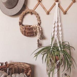 May include: A white macrame plant hanger with a green plant hanging from it. A small woven basket with wooden beads hangs on a wooden wall rack with a gray fedora hat.