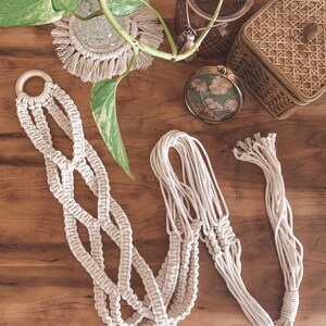 May include: A macrame plant hanger with a wooden ring at the top. The hanger is made of natural cotton cord and has a woven pattern. The hanger is hanging on a wooden surface.
