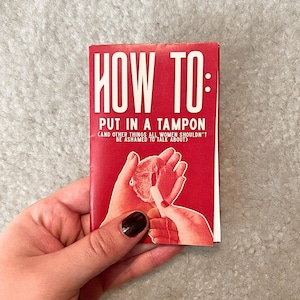 May include: A red booklet titled "HOW TO: PUT IN A TAMPON (AND OTHER THINGS ALL WOMEN SHOULDN'T BE ASHAMED TO TALK ABOUT)". The cover features a graphic of hands and a tampon. The booklet is held by a person.