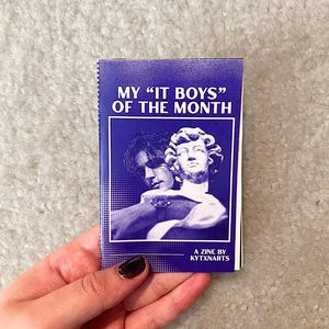 May include: A small, purple zine titled "MY 'IT BOYS' OF THE MONTH" with a black and white image of two figures. The zine is held in a hand with dark nail polish. The text "A ZINE BY KYTXNARTS" is at the bottom.