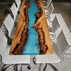 May include: A long wooden table with a blue epoxy river running down the center. The table is surrounded by white office chairs.