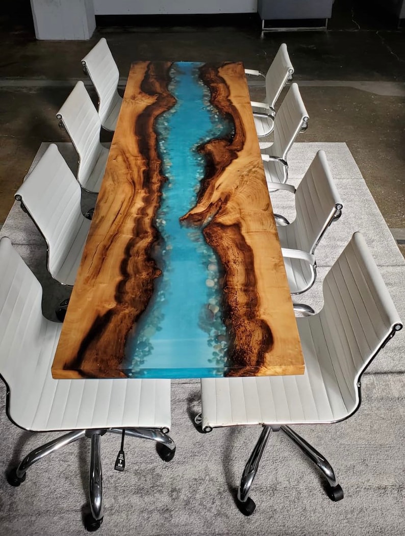 May include: A long wooden table with a blue epoxy river running down the center. The table is surrounded by white office chairs.