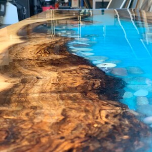 May include: Close-up of a wooden table top with a blue epoxy resin river. The resin has a light blue color and contains white and brown stones. The wood has a dark brown color with a natural wood grain.