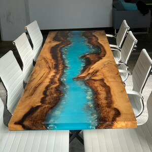 May include: A large wooden table with a blue epoxy river running through the center. The table is surrounded by white chairs.