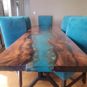 May include: A long wooden dining table with a blue epoxy river inlay and white stones. The table has a black metal base and is surrounded by teal blue velvet chairs.