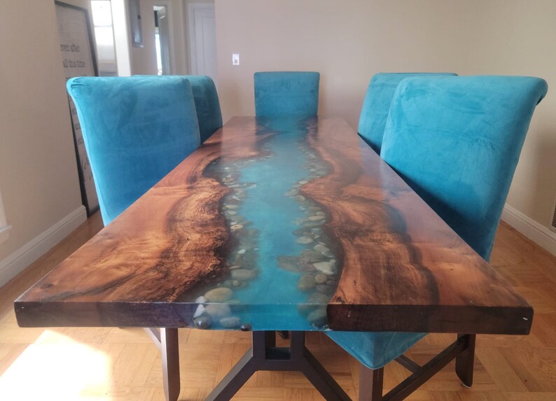 May include: A long wooden dining table with a blue epoxy river inlay and white stones. The table has a black metal base and is surrounded by teal blue velvet chairs.