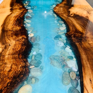 May include: A wooden table top with a river of blue epoxy resin and white and gray stones embedded in the resin.