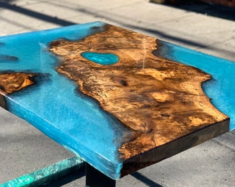 May include: A wooden coffee table with a blue epoxy resin top. The resin has a unique swirl pattern and a small turquoise blue accent. The table has a black metal base.