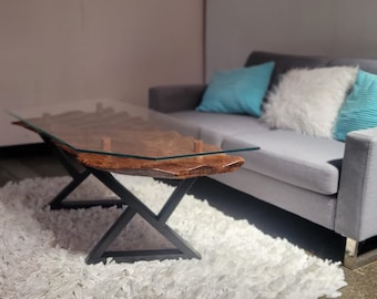 May include: A glass-top coffee table with a wooden base and black metal legs. The table is in front of a gray sofa with blue and white throw pillows.