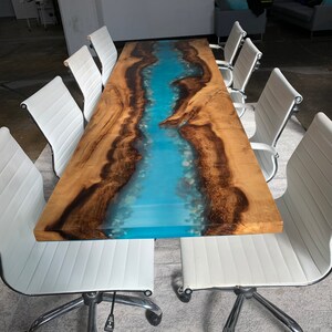 May include: A long wooden table with a blue epoxy river running down the center. The table is surrounded by white office chairs with chrome legs.