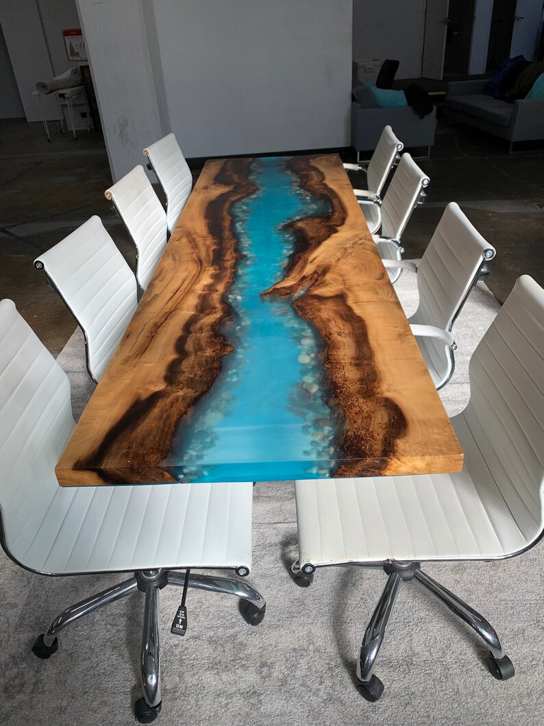 May include: A long wooden table with a blue epoxy river running down the center. The table is surrounded by white office chairs with chrome legs.