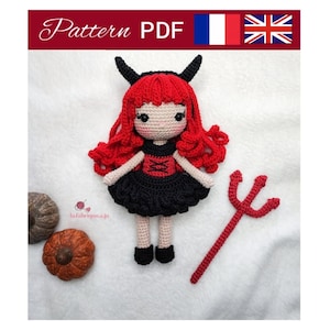 Puede incluir: Una muñeca de diablo de ganchillo con pelo rojo, vestido negro y un tridente rojo. La muñeca tiene cuernos negros y está junto a dos calabazas decorativas. La imagen incluye el texto "Pattern PDF".