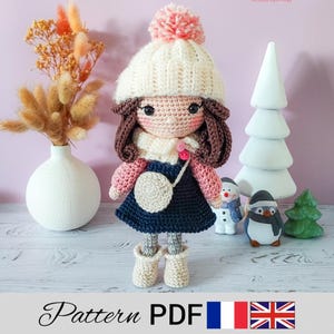 Peut inclure: Patron de poupée au crochet en PDF. La poupée porte un chapeau blanc et rose, un pull rose, une robe bleue et des cheveux bruns. La poupée est debout sur une surface en bois avec un vase blanc et un sapin de Noël blanc en arrière-plan.