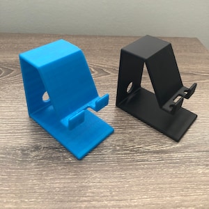 Pode incluir: Dois suportes para telefone, um azul e outro preto, feitos de plástico. Os suportes são projetados para segurar um telefone na posição vertical.