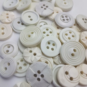 May include: A collection of white plastic buttons in various shapes and sizes. Some buttons have two holes, while others have four holes. Some buttons have a raised design, while others are smooth.